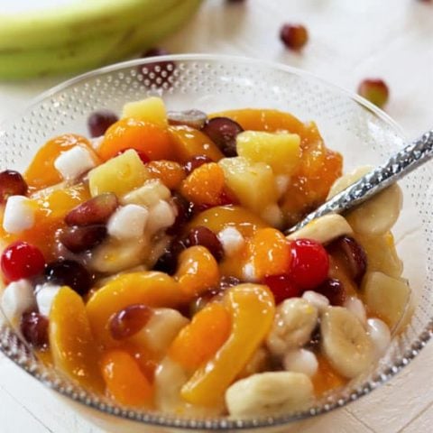 Square image of colorful canned fruit salad recipe in a bowl with peaches, grapes, pineapple, cherries, banana, and marshmallows.